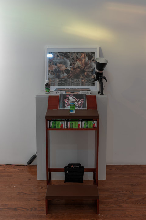 Close up image of a sculpture within the installation featuring a small table in front of a white pedestal. On the pedestal, a picture collage leans against the wall. A ouija board sits in front of the image. On the right, a bottle of Robitussin cough medicine and on the left of the pedestal, a light fixture. In front, on the table, is an image of three men from greco roman period, with massive muscles engaged in what looks like a fight. Another box of Robitussin sits on the table. Below, in a cutout, sit multiple boxes of Robitussin. 