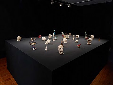 Small ceramic sculptures of skulls, fish, and birds lie on a large black surface against black walls.