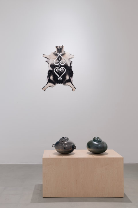 Installation image of works in the exhibition featuring a goat hide on the wall, splayed out with spiral cutouts. It is mounted high above, on the white wall, in the background. In the foreground, two darkly glazed ceramic sculptures sit atop a light brown bench pedestal. 