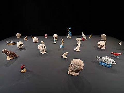 Small ceramic sculptures of skulls, fish, and birds lie on a large black surface against black walls.