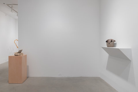 Installation image of two sculptures in the exhibition. On the left, a cement and ceramic sculpture sits atop a light brown wood pedestal. On the right, mounted on a protruding wall shelf, is a stone. Draped over the stone is a piece of metal. 