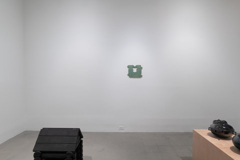 Installation image of a room of sculptures in the exhibition. In the foreground, the roof of a small, black log cabin sits at the bottom of the frame. In the background, an enlarged, light green bread clip, often used to hold bread bags together, is mounted on the wall. On the right, two darkly glazed ceramic sculptures sit atop a light brown wooden bench that is halfway out of the frame. The walls are white and the floor is grey, cement/stone.  
