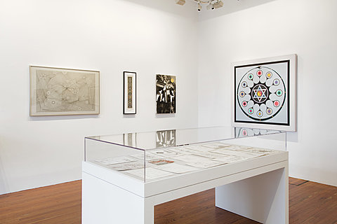 Several framed artworks showing different geometrical diagrams hang on the wall behind a glass display case which holds several sheets of paper.