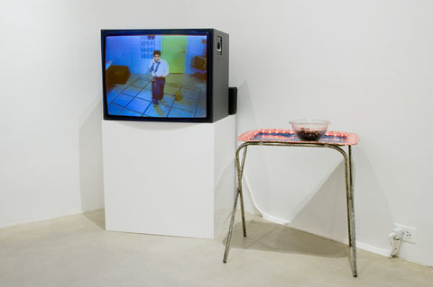 Installation view of the gallery. On the left, a television is on a pedestal featuring a still from a video. The still is mostly green and blue and featuring a single figure in a kitchen-like room. On the right is a small table, with a tray. On the tray is a bowl.   