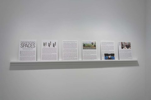 Installation view of six pieces of paper sitting on a white ledge against a white wall. The pieces of paper are in black and white and contain black typed text, but the words are illegible from the camera view point and image quality. 