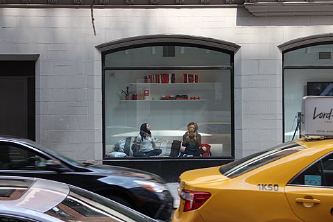 Two people sit in a window display in front of a bookshelf. A car and a taxi pass by in the foreground.