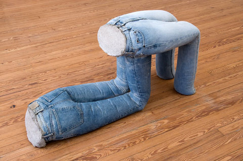 Close-up image of a sculpture featuring two pairs of jeans filled with cement. The first pair is front facing on the ground, bent at the knees holding the other pair up by its buttocks. The second pair of jeans also has its legs bent at the knee. 