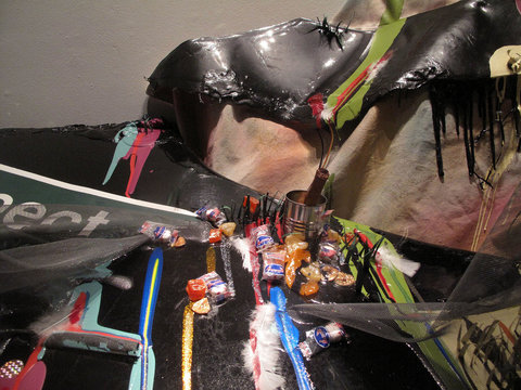 Close-up image of the window installation featuring a pile of what could be perceived as trash. Included is a street sign, a bottle, a can, some wrappers/snacks, and a bunch of random materials placed in a seemingly random pile. 