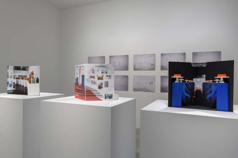 Installation view of three pedestals in the exhibition. On the left side, a white pedestal with a pamphlet on top. In the middle, a white pedestal with another white pamphlet on top and various illegible sketches. On the right, a white pedestal with a black and blue pamphlet on top. In the background, several black and white images are mounted to the wall in two rows of five. 