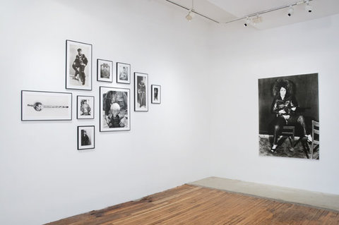 Installation view of the gallery featuring two walls. On the right wall is a collection of nine framed works. On the right wall is a black and white painting of a woman with an afro, sitting on a chair. 