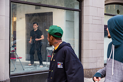A person dances in a window display; two passers-by on the street look on.