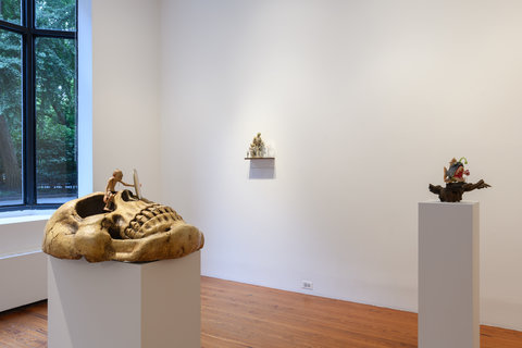 Gallery installation view featuring a pedestal in the left foreground, a pedestal on the right middle ground, and a work on the back wall. On the pedestal in the foreground, there is a large skull facing the ceiling. Atop the skull sits a small Gollum figurine, a character from Tolkien that is small and skinny with goblin-like features. It holds onto a small circular object and peers into it. It is naked and has string like hair and wide eyes. On the pedestal in the middle ground is an angler fish with a wide open mouth revealing long and sharp white teeth and a green antenna dangling above a fish just in front of its mouth. The fish is an orange clown fish and the scene is supposed to resemble Pixar's FINDING NEMO animated movie. Both fish figurines are on top of a thick, dark brown branch. In the back of the view, on the wall is a shelf. On top of the small shelf area a group of zombie figurines in various sizes. To the left in the back is the gallery window which is open, allowing for sunlight and a view into the park across the street which is fraught with green foliage.