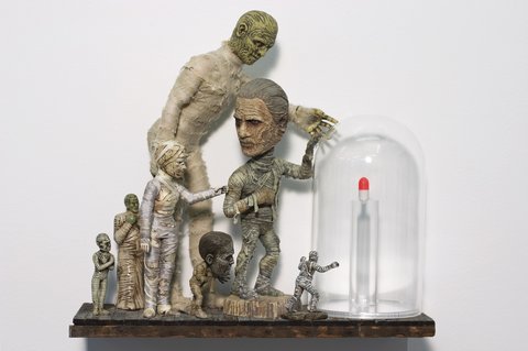 Image of a sculpture featuring several zombie and mummy figurines jumbled together in various sizes. On the right of the sculpture is a white and red pill encased in a glass half-cylinder. 