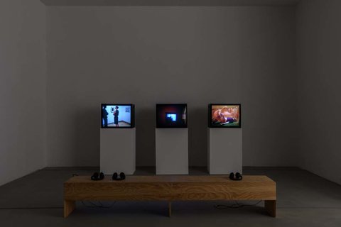 Installation view of a dark room featuring three television monitors in a row perched on three white pedestals. In front of the monitors, sits a light wood colored bench. 