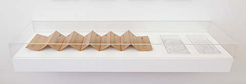 A glass display case showing an accordion-folded scroll of paper and two printed sheets.
