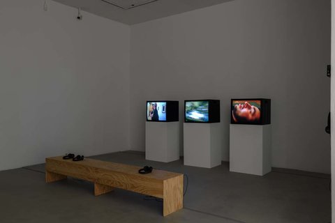 Installation view of a dark room featuring three television monitors in a row perched on three white pedestals. In front of the monitors, sits a light wood colored bench. 