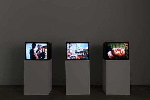 Installation view of a dark room featuring three television monitors in a row perched on three white pedestals.