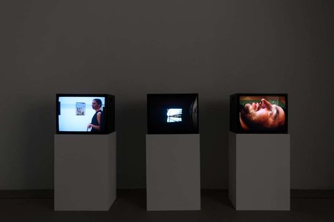 Installation view of a dark room featuring three television monitors in a row perched on three white pedestals.