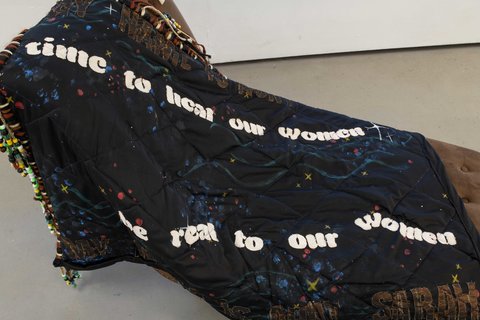 Close-up view of the sculpture in the exhibition. It appears to be a dark brown lounger chair covered by a large black piece of textile with beads and the text “time to heal our women”, “be real to our women”, and “MY AUNT IS AUNT SARAH”.