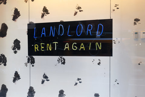 A close up view of the interior of a window display. In the center a black sign with neon blue and yellow lights reads: Landlord rent again. Landlord is in blue and rent again is in yellow. Behind the sign is a black and white spotted background.  