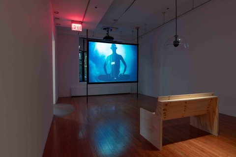 Installation view of the video installation in the gallery space. The room consists of one large space with a large projector a the end. On the left, a doorway lets in light to another room, on the right, a white wall sits in the shadows. In the foreground of the exhibition, a wooden bench sits facing the projection. 