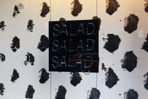A close up view of the interior of a window display. In the center a black sign with neon blue lights reads: Salad, Salad, Salad. Behind the sign is a black and white spotted background.  