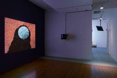 Installation view of a room in the gallery. On the left, a projector screen is hung against a dark wall. The lights are off and the source of light comes from the screen. The still from the video consists of a black figure with a white ball and black hood where their head is supposed to be. The background is orange. In the middle, a powered-off television monitor hangs on the wall. On the right, a hallway leads to another room in the gallery. 