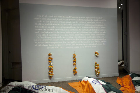 Installation view of the exhibition featuring wall text above three segments of hanging orange and white flowers on the wall. The text is unable to be discerned from the camera's point of view. In front of the wall, there are three fake bodies covered in flags that are stitched together down the middle. The flags are half the flag of India and half the flag of Pakistan. While we know these are fake bodies, they are only partially in view from the camera's point of view. 
