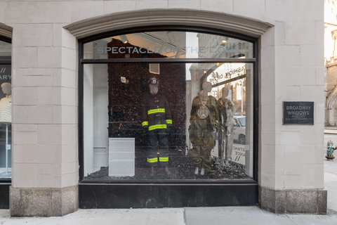 Image of two mannequins in the exhibition. The mannequin on the left wears a black and yellow fire-fighter looking outfit, while the mannequin on the right is draped in brown fabrics. On the very far left, is a white square with exhibition text, but it is illegible from the camera view.  