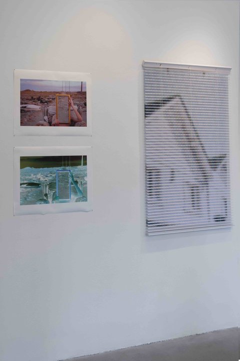 Installation view of three works in the exhibition. On the left, two smaller works are hung above the other. The small image on top consists of a figure seated in a rocky, outdoor pasture holding a mirror over their body so that there is no face or torso. The image below is a negative of this image. On the right a close up image is very blurred but seems to be a composition of a photograph taken of window blinds. 