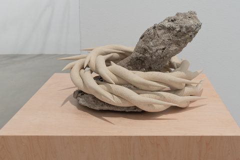 Installation image of a sculpture in the exhibition sitting atop a light brown wood pedestal. The sculpture features a piece of rugged cement/rock wrapped with a coil of spiky ceramic rings. 