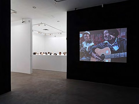 A projection on two people playing acoustic guitars is on a black wall. In the background is a room with white walls and multiple sculptures.