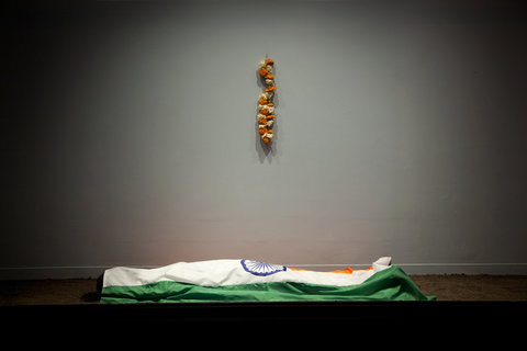 Installation image of the exhibition featuring orange and white flowers, hung on the wall in a line. Below is a folded flag with a fake body underneath it. The flag, while only part is shown in the camera's point of view, the green and white colors and symbol show that it is part of India's flag. 