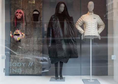 Image of two mannequins in the exhibition. On the right side of the image, an ornate fabric is draped and three mannequin heads with hats and masks are mounted on it. In the middle, a mannequin wears a very puffy black coat and black tights. On the right, a mannequin wears a light, white, puffer jacket with its hands on its hips. The mannequin on the right does not have legs.