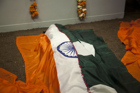 Close-up view of the installation featuring the bottom of a wall and the installation on the ground in front of it. On the wall, there are two strings of orange and white flowers. Below, is a fake body covered with a flag. The flag is half the flag of India and half the flag of Pakistan. It features the orange and white side of the India flag and half of the crescent moon of the Pakistan flag which is dark green with a white crescent. 