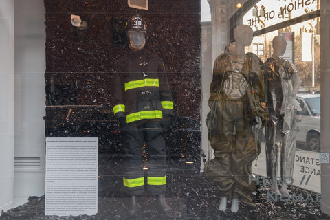 Image of two mannequins in the exhibition. The mannequin on the left wears a black and yellow fire-fighter looking outfit, while the mannequin on the right is draped in brown fabrics. On the very far left, is a white square with exhibition text, but it is illegible from the camera view.  
