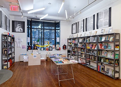A table in the middle of the room is stacked with colorful magazines and printed media. The two parallel walls to the table have black industrial bookshelves filled with printed media. Above the bookshelves are large posters which consist of black and white printed texts. The wall perpendicular to the table features a large bay window with printed papers stuck to the bottom half of it. 