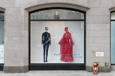Image of two costumes from the exhibition on mannequins. On the left, a mannequin wears a black wetsuit with red stripes along side the body. It also wears goggles and a swim cap. Next to it, on the right, another mannequin wears an ornate red dress with a red bonnet-like hat. The image is from the street view. 