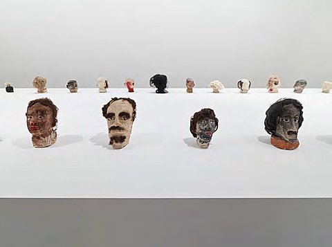 A large collection of small sculpted heads, all facing outward, are displayed on a massive white platform.
