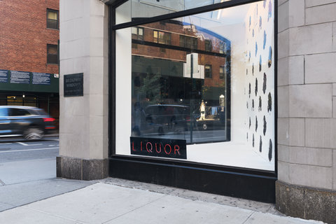 Corner view of a light colored building with a large window. In the window is a small black sign on the bottom left corner. The sign reads the word liquor in red neon lights.