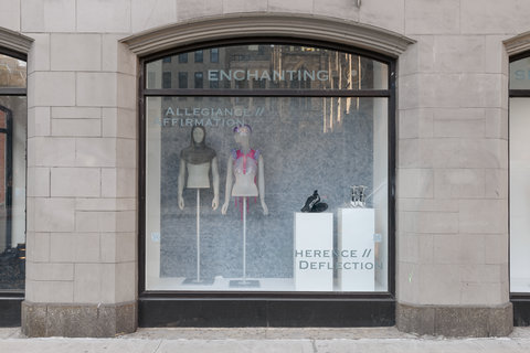 Image of two mannequins in the exhibition and two pairs of shoes. On the left, a mannequin wears a grey hood, and nothing else. The mannequin is missing the lower half of the body. Next to this mannequin, another mannequin wears a pink and purple crocheted item around their head and shoulders. In the foreground, through the window, a pair of black sneakers sits on a pedestal next to a pair of white heels with silver spikes along the straps. The image is taken from the street view. 