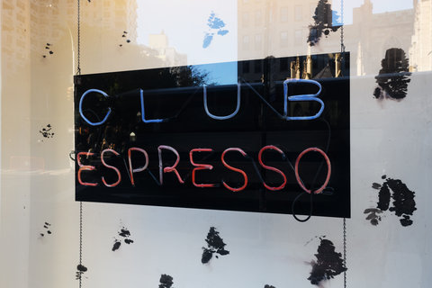 A close up view of the interior of a window display. In the center a black sign with neon blue and red lights reads: club espresso. Behind the sign is a black and white spotted background.  