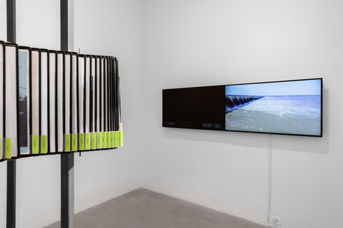 Installation view of the exhibition featuring flippable archival papers and articles surrounding two poles. On the left, two television monitors are mounted to a white wall. 