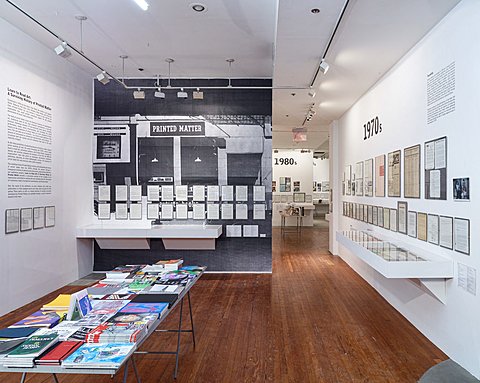A table in the middle of the room is stacked with colorful magazines and printed media. The half wall perpendicular of the table features a black and white image with printed out framed texts over it. The right parallel wall is lined with a series of printed matter from the 1970s. The left parallel wall features more text about printing media in the 1970s. Through the hallway there is a glimpse of the following room that follows printing media in the 1980s.
