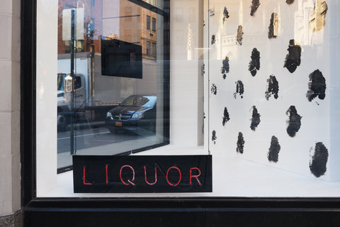 A close up view of the interior of a window display. In the lower left corner of the window a black sign with neon red lights reads: liquor. Behind the sign is a black and white spotted background.  