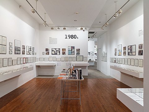 The room showcases printed media history from the 1980s. There is a table in the middle of the floor that has magazines, books, and other printed media stacked on top of it. A series of framed printed texts from the 1980s is lined up on each wall. There is a table top mounted on each wall underneath the framed texts featuring more printed media laid on its surface.