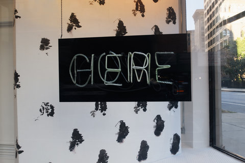 A close up view of the interior of a window display. In the center a black sign with neon blue lights reads: come here. The two words lay over one another. Behind the sign is a black and white spotted background.  