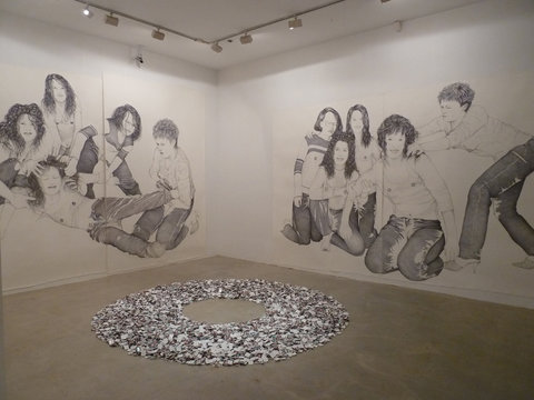 Gallery installation view of two large ballpoint pen drawings. On the right is a wall-sized drawing of many of four figures intertwined, engaging with one another and touching one another. On the left, there is a wall-sized drawing of four figures kneeling in a group while another figure approaches them. In front of the works, is a pile of matchboxes, arranged in a large circle with a hole in the middle. 