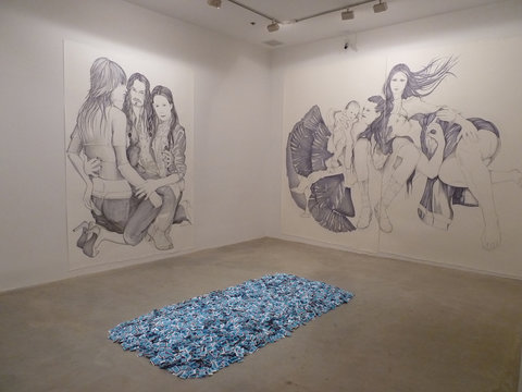 Gallery installation image of two large ballpoint pen drawings. On the right is a wall-sized, floor to ceiling, image of three kneeling figures intertwined with one another. On the left is a wall-sized ballpoint pen drawing of intertwining figures with swirling hair. On the floor in front of the work is a pile of blue matchboxes arranged in a long rectangle. 