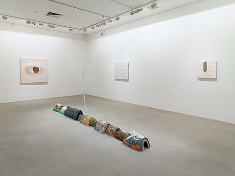 A line of colorful small tunnel-like shapes is arranged on the floor of the gallery. Three mildly colorful images hang on the wall behind the floor sculpture.